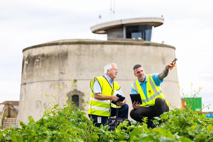 Two male Mitie engineers wearing high vis vests, crouching in front of Martello Tower in Essex. The man on the right-hand side is pointing off-screen with a tool in his hand