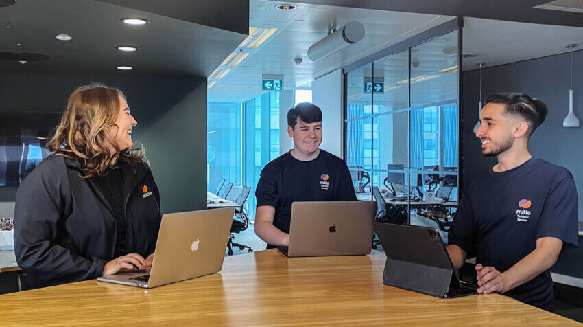 Three young people in Mitie apprentice-branded t-shirts laughing with each other. They stand around a wooden table in an office, using laptops.