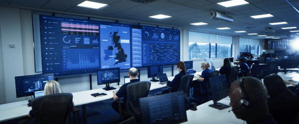 Mitie TSOC security centre - people sitting at rows of desks with computers, with a large screen of data in the UK showing on the far wall