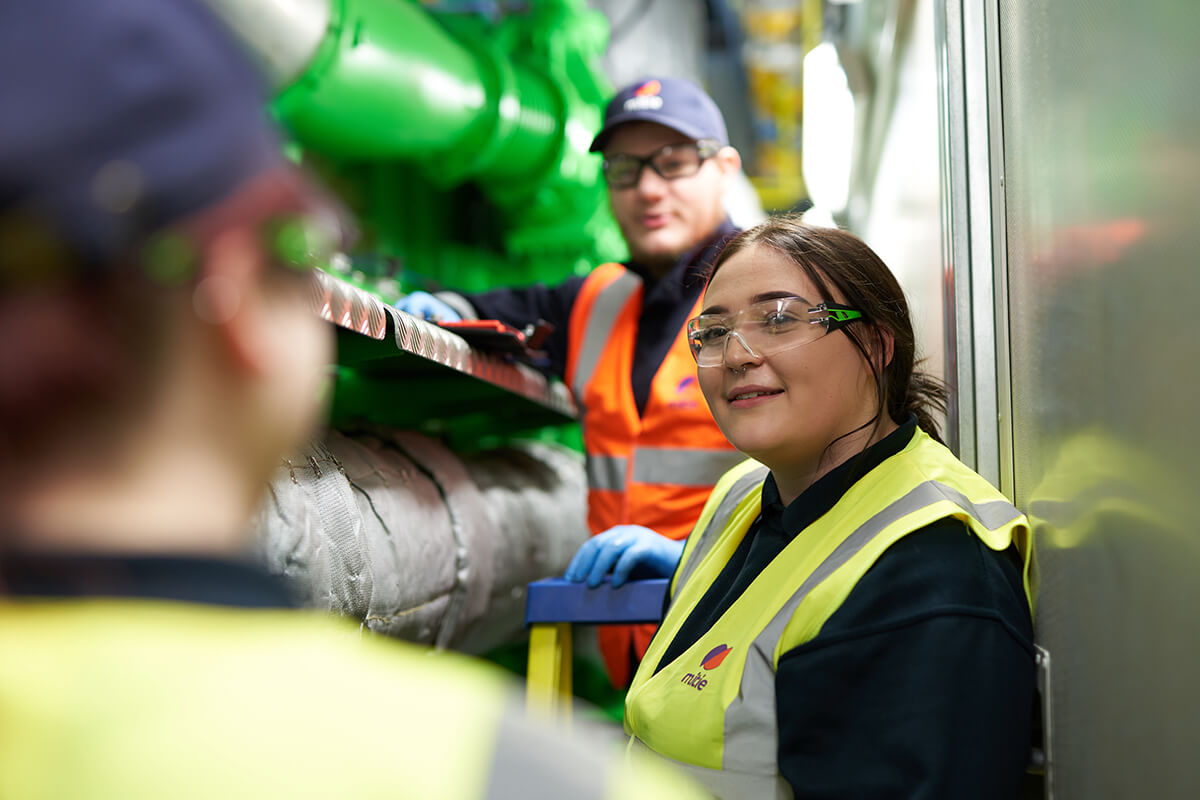 Female engineer in Mitie branded hi vis and safety glasses, standing next to large pipes