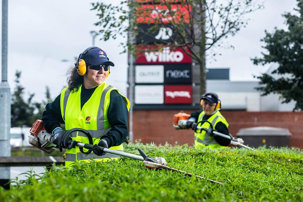 Two women wearing Mitie-branded high vis jackets, hats and safety glasses, strimming the top of a hedge