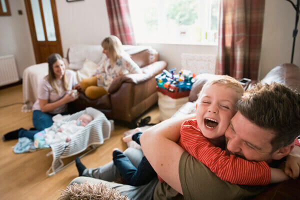 A happy family in a living room; foreground, a laughing boy hugs his father