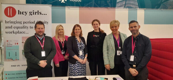 Mitie staff members at a Hey Girls stand - the pull-up banner behind says 'Bringing period dignity and equality to your workplace'