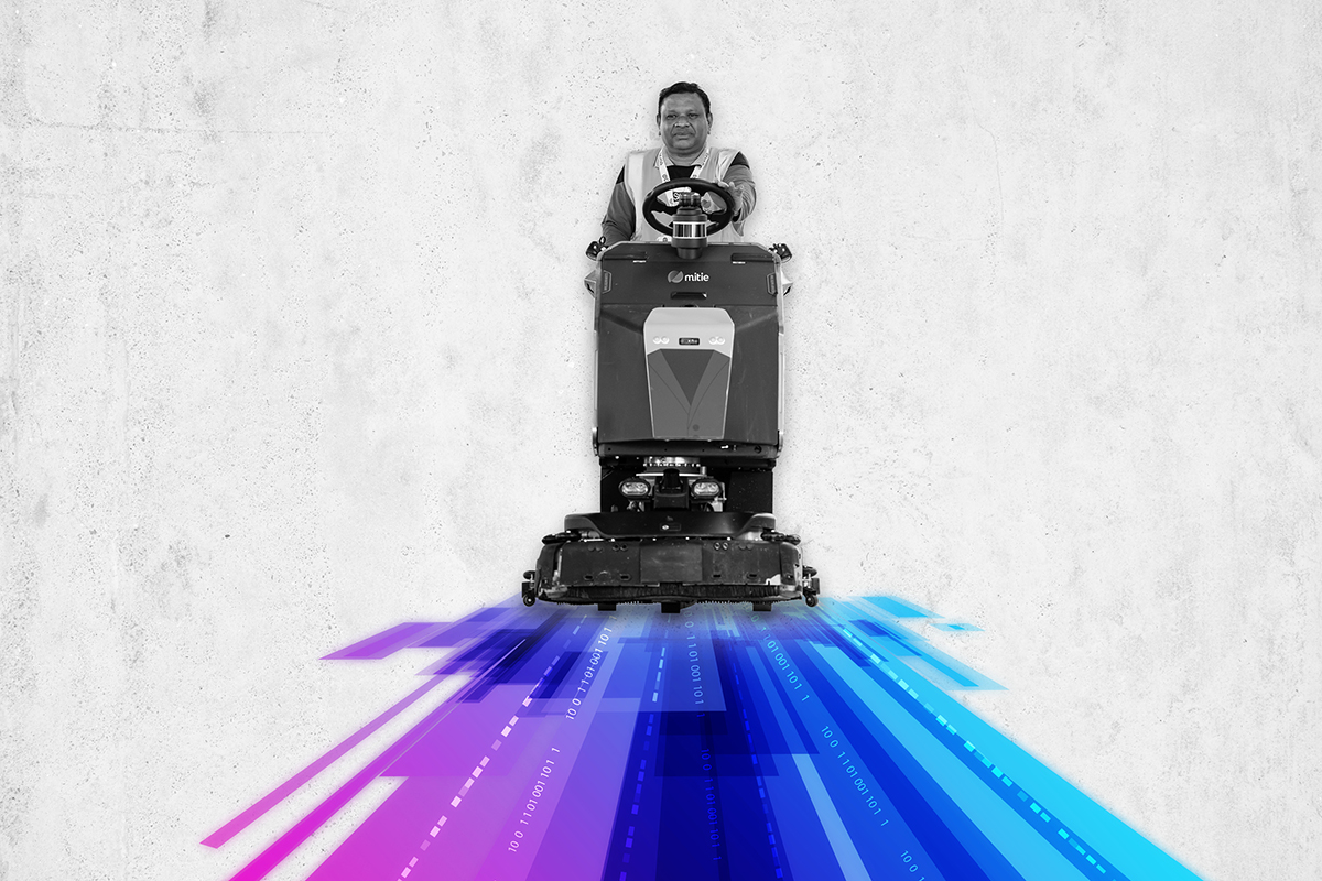 A Mitie colleague in black and white on a ride-on cleaner, with stripes of purple, blue and magenta underneath them