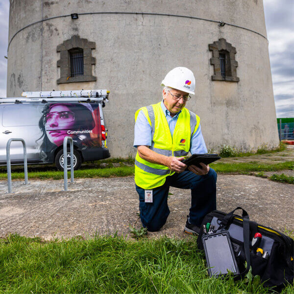 A man in Mitie hard hat and high vis kneeling in front of a concrete building and using a digital tablet