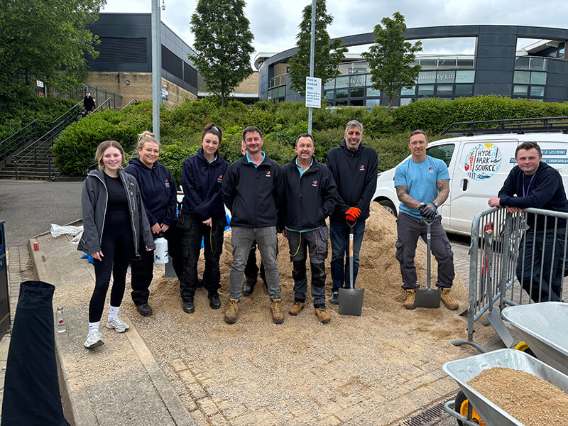 A group of smiling Mitie colleagues, standing in front of a pile of gravel and holding spades